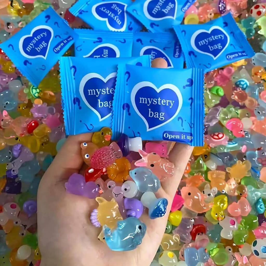 A hand holds colorful animal-shaped charms above a surface covered in more charms and several blue "mystery bag" packets with question marks and a heart design.