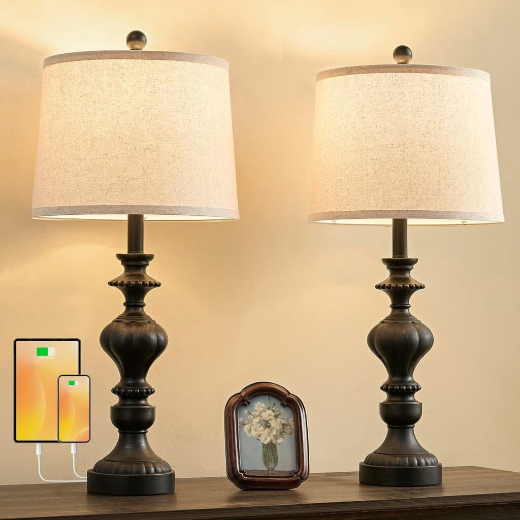 Two dark bronze farmhouse table lamps with beige shades sit on a wooden surface, adding a touch of farmhouse decor to any room. Between them is a small framed floral photo. A phone graphic highlights the convenient charging feature via USB ports on this table lamps set.