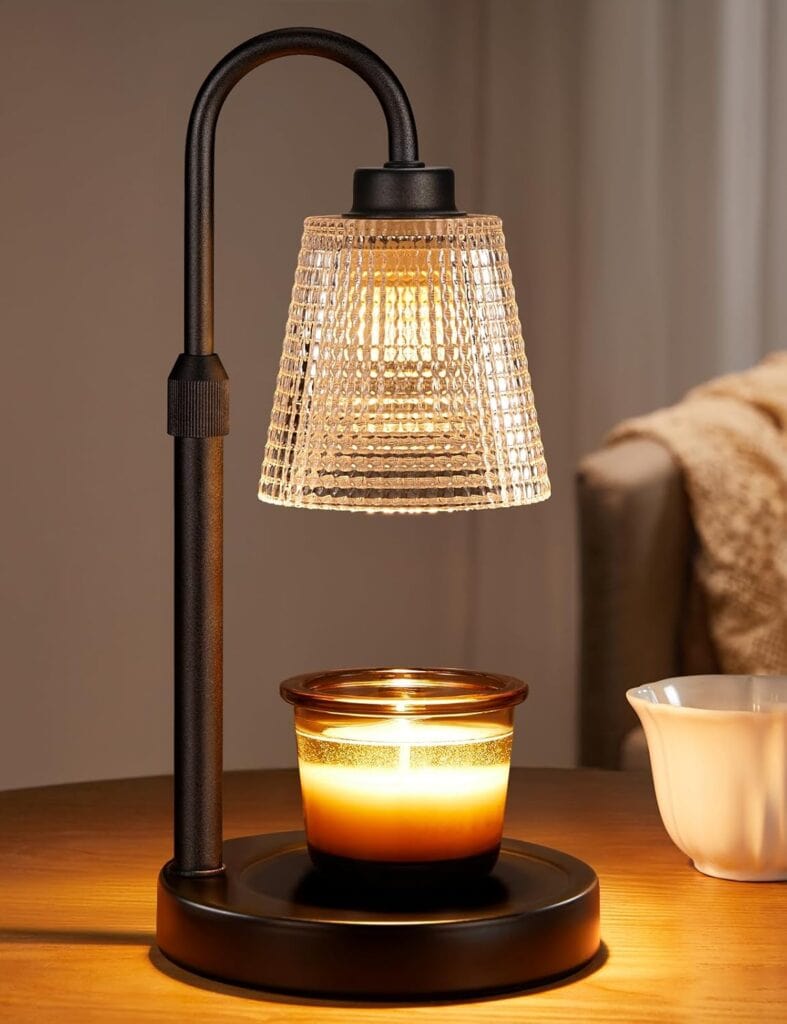 A candle warmer lamp with a ribbed glass shade shines light onto a lit jar candle on a wooden table, next to a white dish and a cozy beige blanket in the background.