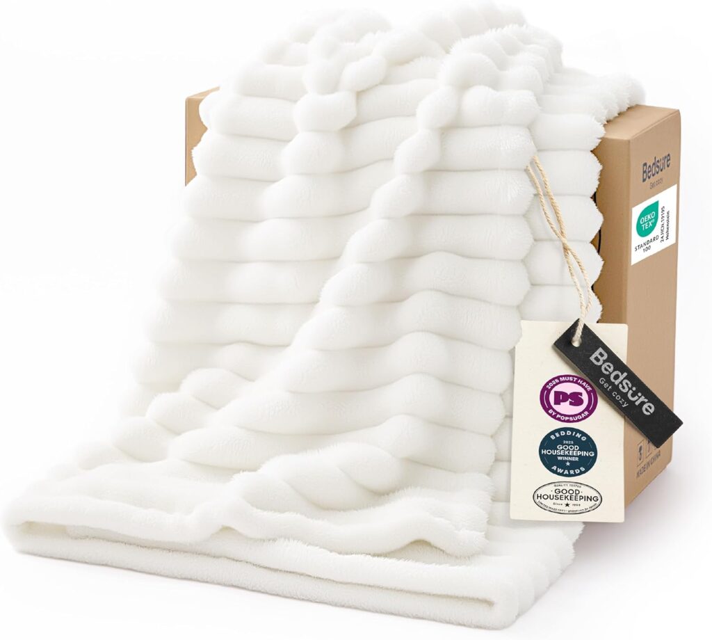 A white, textured fleece blanket draped over a brown box, with product tags attached. The blanket features a ribbed design and looks soft and plush.