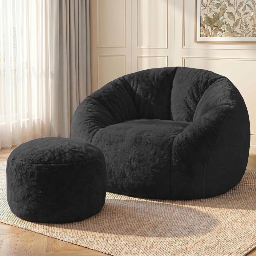 A plush black bean bag chair with a matching round ottoman sits on a woven rug in a bright room with light walls, large windows, sheer curtains, and framed botanical artwork.