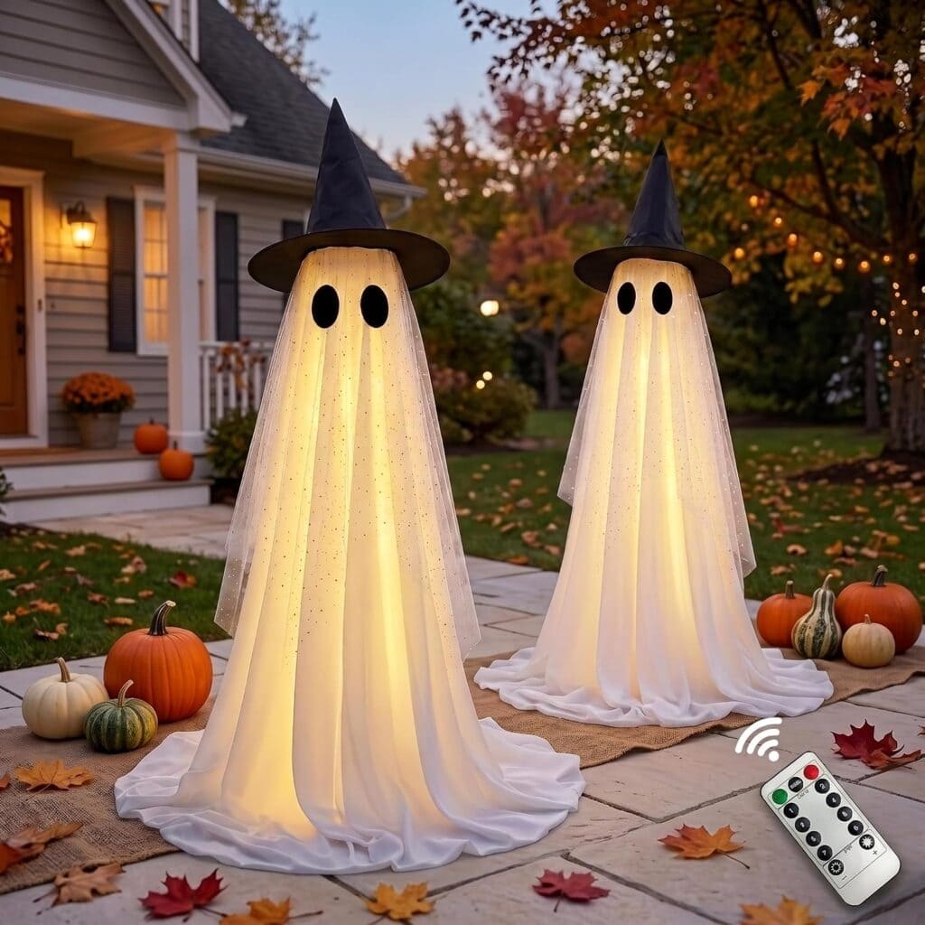 Two glowing ghost decorations with witch hats stand on a porch surrounded by pumpkins and autumn leaves. A remote control is visible in the corner. The scene is set during fall by a house with a lit porch.