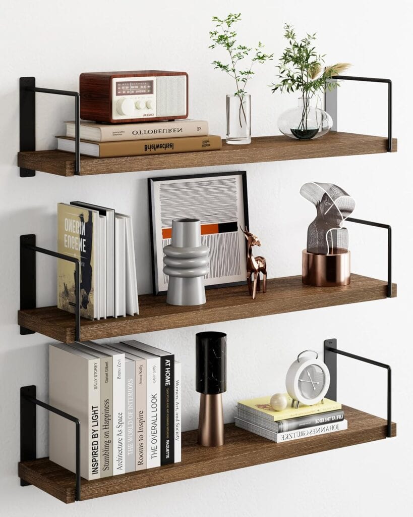 Three wooden wall shelves hold books, decorative objects, plants, a small radio, and a clock against a white wall. Featuring black metal brackets and modern decor items, these shelves are perfect for updating your space—especially with our exclusive Friday Deals.