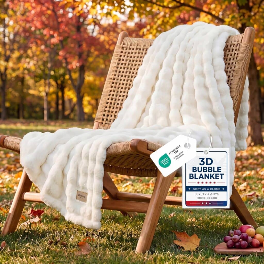 A white, textured 3D bubble blanket draped over a woven wooden chair sits outdoors on grass, with autumn trees in the background. Product tags are attached to the blanket, and grapes rest on the ground nearby.