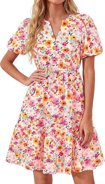Woman wearing a knee-length, short-sleeve, pink floral dress with a v-neck and gathered waist. The dress features a colorful pattern with orange, yellow, and pink flowers.