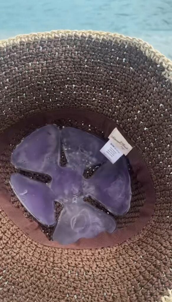 A view inside a brown woven hat with a translucent purple sand dollar and a white tag, perfect for pairing with a cooling hat insert to help keep you cool. Out-of-focus blue water shimmers in the background.