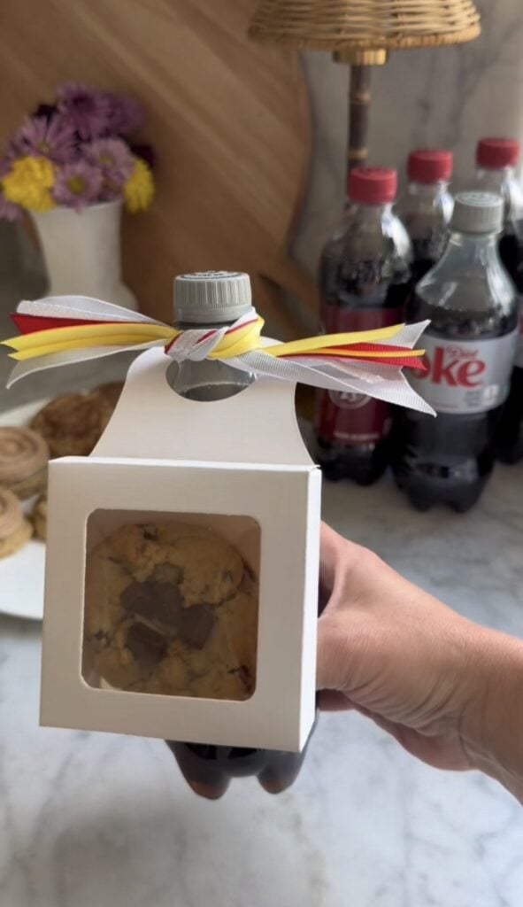 A hand holds a Diet Coke bottle with a white box containing a cookie attached to its neck, decorated with yellow, white, and red ribbon—an affordable teacher gift. More Diet Coke bottles and flowers are visible in the background.