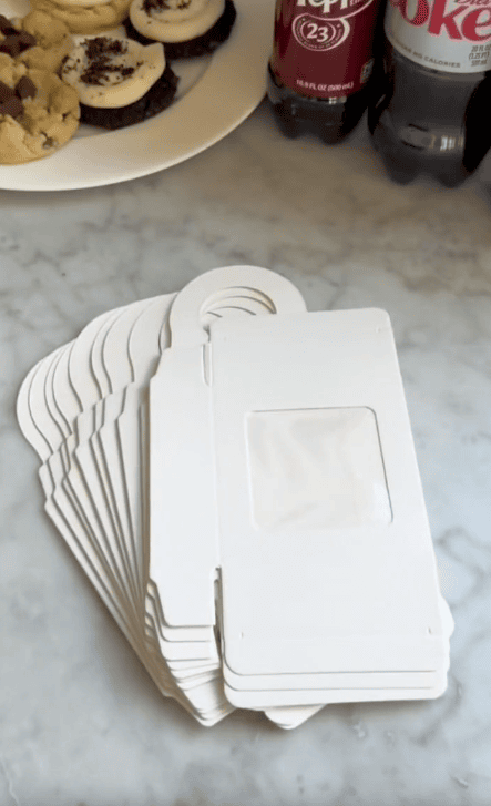 A stack of blank white door hangers with rectangular cut-out windows lies on a marble countertop—perfect for an easy DIY teacher gift. Nearby are plates of cookies, sliced bananas, and bottles of soda, including Diet Coke and Dr Pepper.
