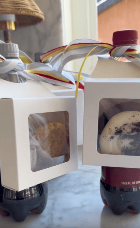 Two small white boxes with clear windows, each containing baked goods, are tied with colorful ribbon and placed on soda bottles—a charming and affordable teacher gift arranged on a marble surface near a window.