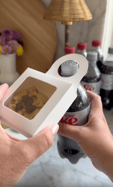 A person holds a bottle of Diet Coke in one hand and a small white box with a cookie inside in the other—an easy teacher gift idea. Several more Diet Coke bottles are visible on the counter in the background.