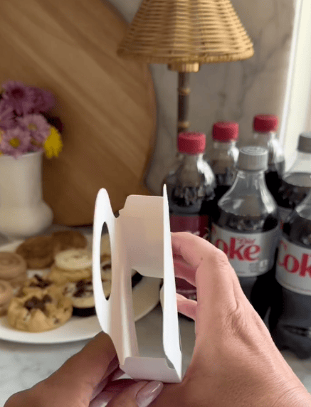 A hand holds a white cardboard drink carrier—an easy teacher gift—in front of a table with Diet Coke bottles, assorted cookies, flowers in a vase, and a wooden cutting board.