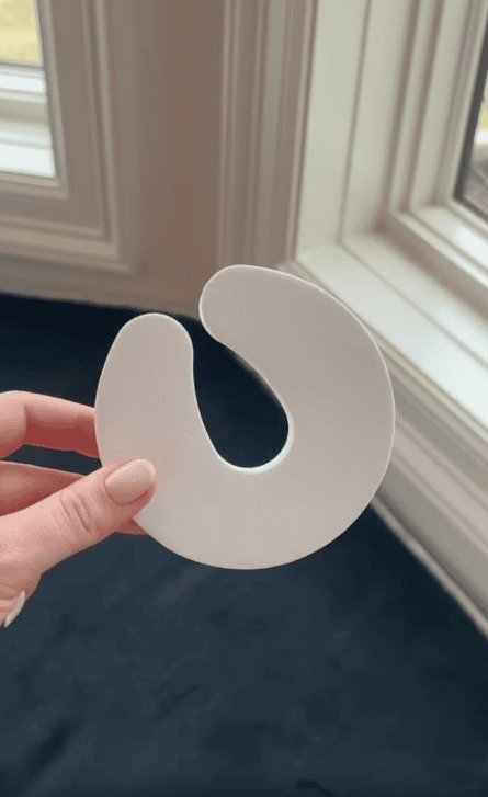 A hand with pale nail polish holds a white, crescent-shaped door pinch guard—perfect for childproofing and enhancing kids safety—in front of a window with white trim and a dark floor.
