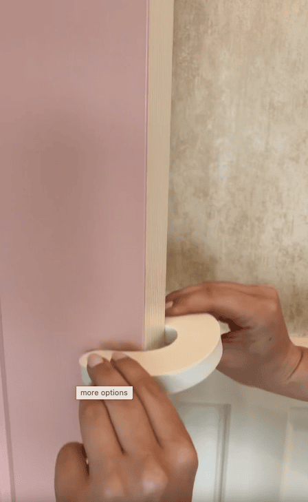 A person’s hands apply white adhesive tape to the edge of a pink door, using a door pinch guard for childproofing, against a textured beige background. A “more options” label appears near the tape.