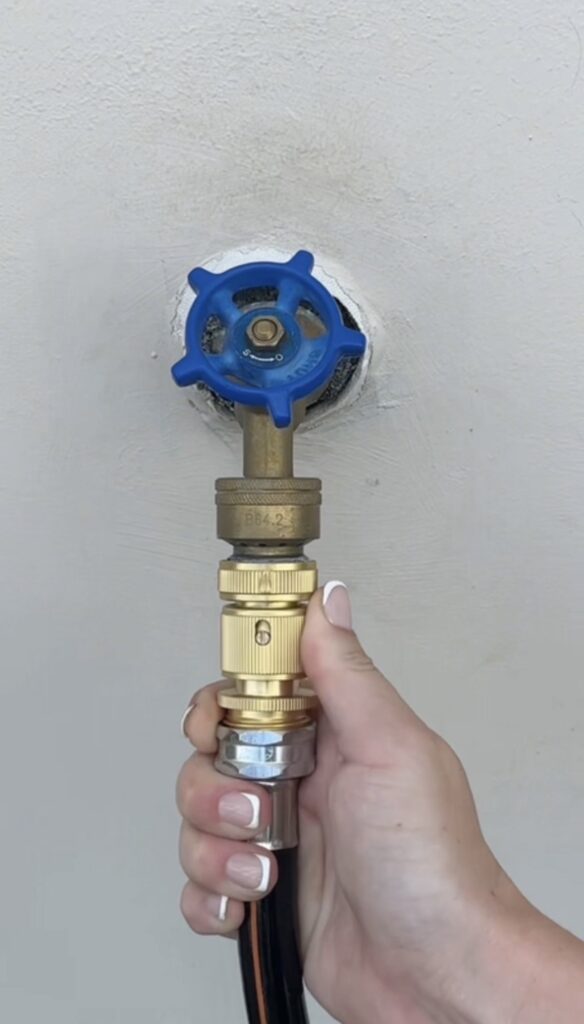 A hand holds a brass garden hose connector attached to a black hose, connecting it to an outdoor blue-handled water spigot—showcasing convenient Garden Hose Quick Connect garden hose accessories.