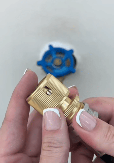 A person with manicured nails holds a brass garden hose quick connect fitting in front of a blurred blue valve attached to a white surface.