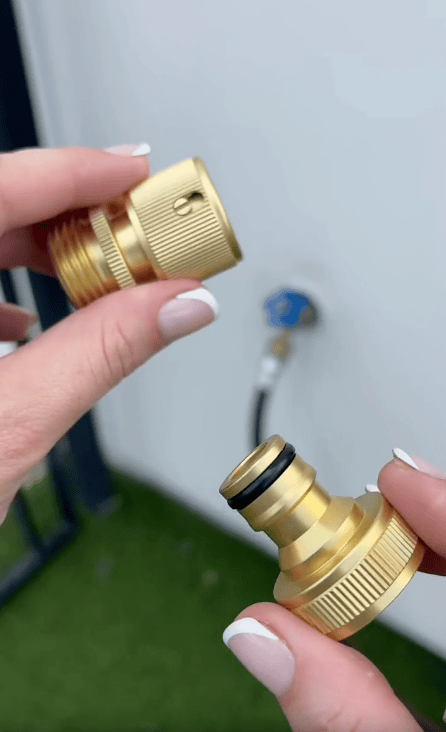 A person with light-colored nail polish holds two brass Garden Hose Quick Connect fittings near an outdoor faucet. The background shows a white wall, blue faucet, and green artificial grass.