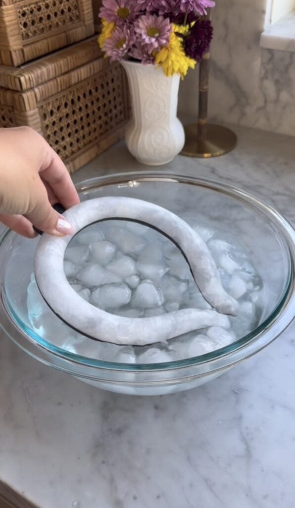 A hand holds a flexible, U-shaped neck cooling tube above a glass bowl filled with water and ice cubes, on a marble countertop with flowers and woven baskets in the background—definitely worth it according to any mom review.