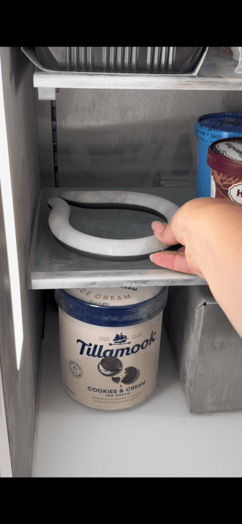 A hand slides out a frosty freezer shelf, holding a round slab of ice above a container of Tillamook Cookies & Cream ice cream—perfect for chilling your Neck Cooling Tube and keeping other frozen items fresh.