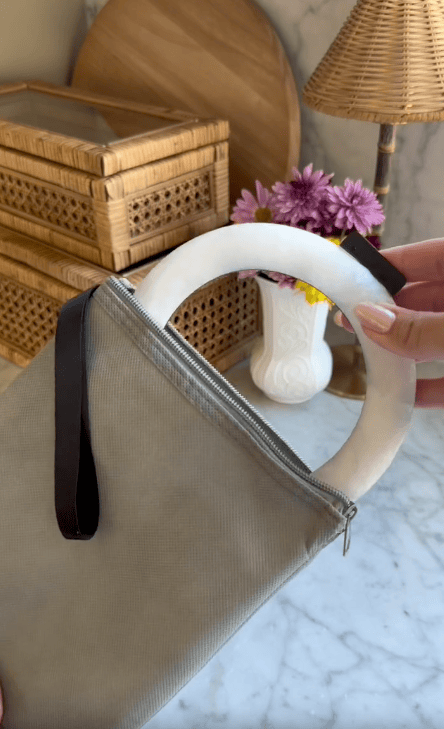 A hand holding a gray zippered handbag with a circular white handle, next to a white vase of purple flowers and wicker boxes on a marble surface—perfect for any mom review.