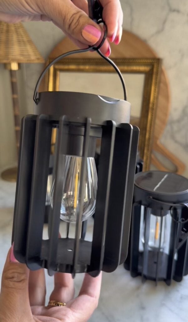 A hand with pink-painted nails holds a cozy black Solar Hanging Lantern with a visible LED bulb; another lantern and backyard decor items are softly blurred in the background.