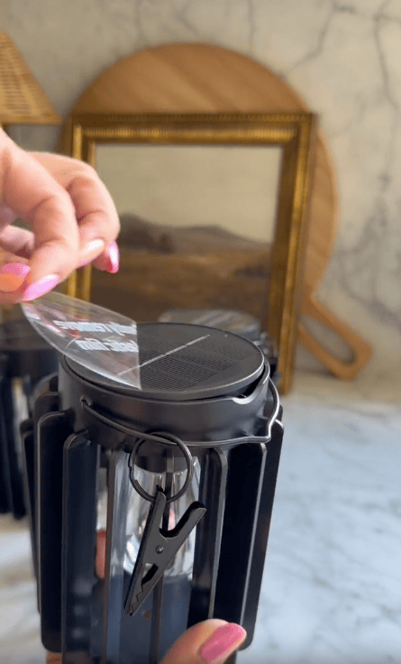 A hand with pink nails peels a protective film off the solar panel atop a black outdoor Solar Hanging Lantern, adding a cozy touch to the backyard. Wooden frames and a cutting board create a warm, blurry background.