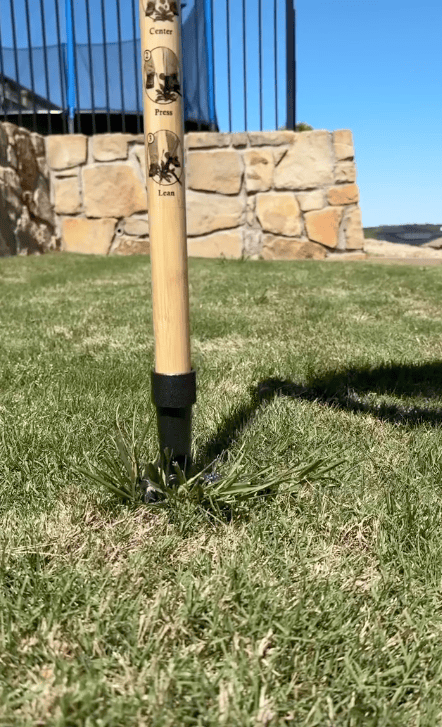 A Stand Up Weed Puller is inserted into a patch of grass on a lawn, with a stone wall and blue sky in the background. The tool's instructions—Center, Press, Lean—are visible, making weed removal easier than pulling weeds by hand.