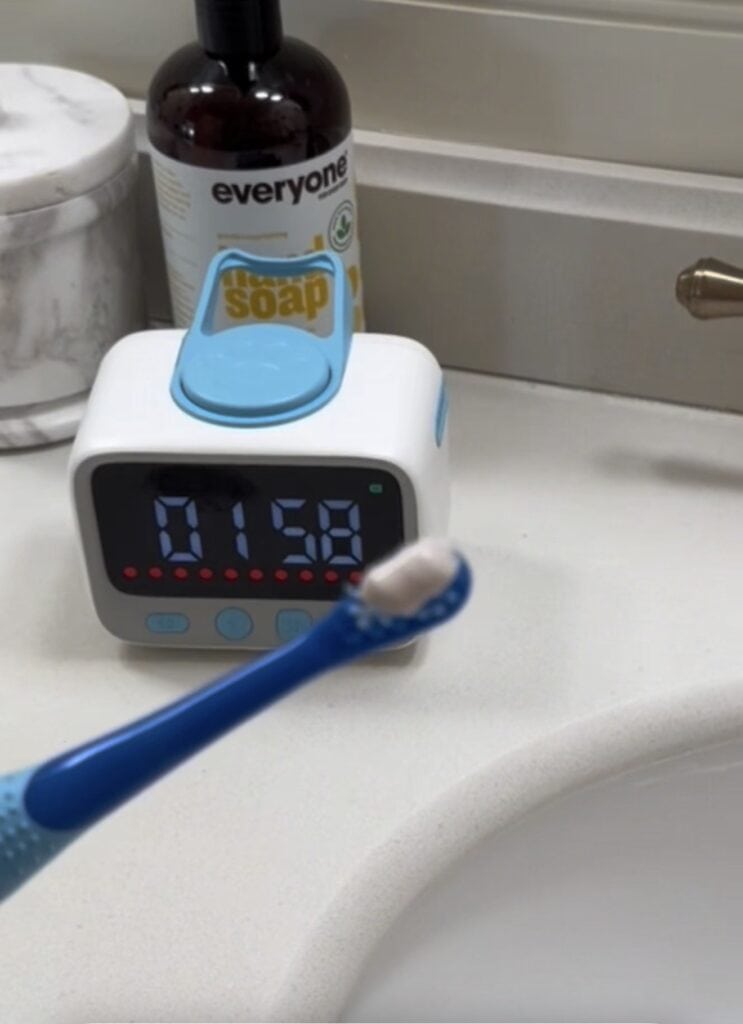 A blue toothbrush with toothpaste is held in front of a bathroom sink. In the background, a Visual Timer for Kids reads "01:58," helping to reduce daily battles. A soap dispenser and a bottle labeled "everyone" are also visible.