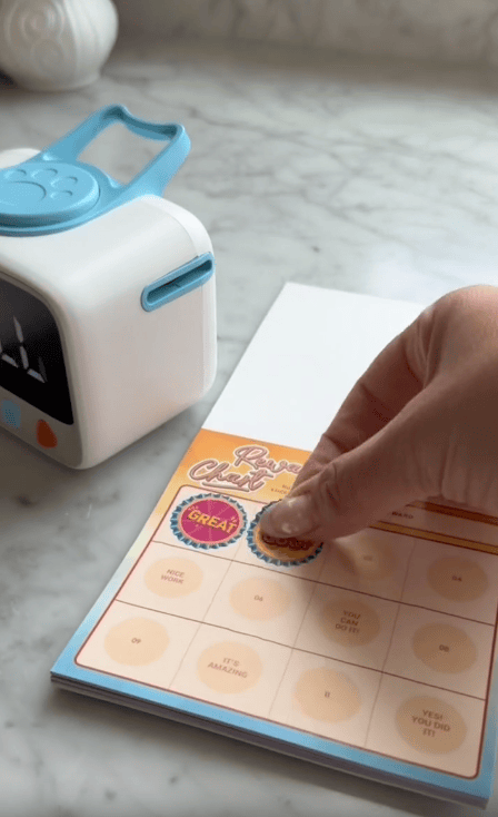 A hand places a colorful sticker labeled “GOOD” on a reward chart filled with praise phrases. Next to the chart, a Visual Timer for Kids sits on the marble surface, helping to reduce daily battles and encourage positive behavior.