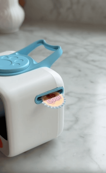 A small white and blue printer sits on a marble countertop, next to a kid-friendly Visual Timer for Kids, printing out a colorful circular sticker that says "So Cool.
