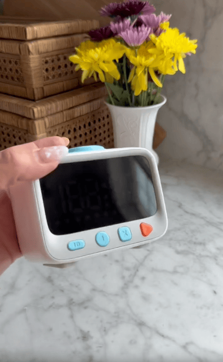A hand holds a small visual timer for kids with blue and orange buttons on a marble countertop. Behind it, wicker boxes and a white vase with yellow and purple flowers help create an inviting space to reduce daily battles.