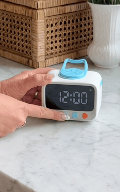 A hand points to a small digital clock displaying 12:00, featuring blue and orange buttons for a visual timer for kids. The clock with its blue handle sits on a marble countertop near a wicker basket and white vase—perfect for easing daily routines.