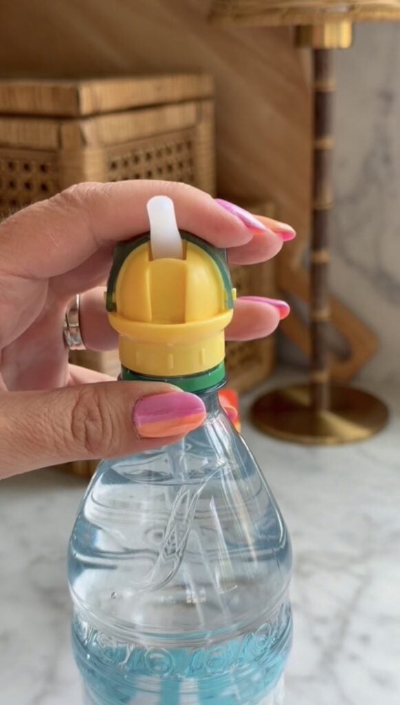 A hand with pink and orange nails holds a water bottle with a yellow and green straw cap. The background includes wicker boxes, a brass lamp, and a marble surface.