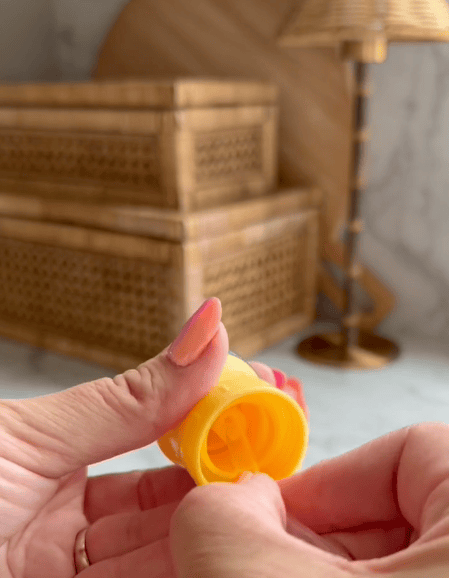 A person with coral-painted nails is holding and opening a small yellow plastic capsule, its shape reminiscent of a water bottle cap. Blurred wicker boxes and a lamp are in the background.