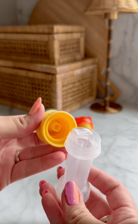 A person with pink and orange nails holds a small transparent plastic container and its yellow water bottle cap, with straw wicker boxes and a lamp in the background.