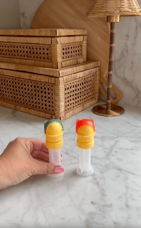 A hand holds two plastic popsicle molds with yellow lids, resembling a water bottle cap with straw—one contains a red popsicle, the other green. Woven boxes, a wooden cutting board, and a lamp sit on a marble countertop in the background.