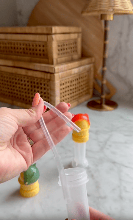 A hand holds a clear plastic straw above a container, with woven baskets, a water bottle cap, colorful bottle caps, and a lamp in the blurred background on a marble surface.