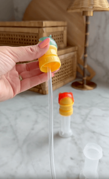 A hand holds a yellow and teal bubble wand with a long clear bubble film. Nearby, another bubble container with a red lid—resembling a water bottle cap—stands on a marble countertop, with wicker boxes and a lamp in the background.