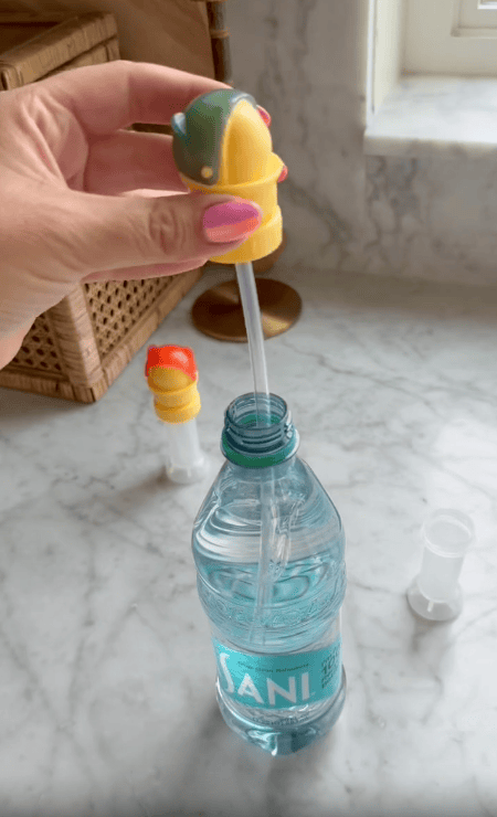 A hand holds a dolphin-shaped water bottle cap with straw above a water bottle on a marble counter, with other colorful dispensers and caps nearby.