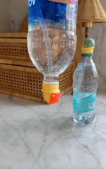 A plastic water bottle with an upside-down straw cap from a sports drink is held above a table, next to another bottle with a yellow and green cap. Wicker drawers and a lamp are in the background.