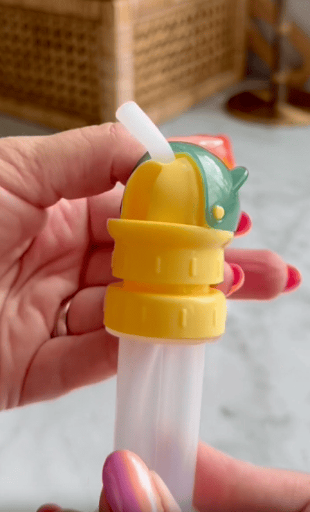 A hand holds a yellow and green dolphin-shaped water bottle cap with a white straw attached to a clear plastic tube. The person has pink and orange painted nails. The indoor background shows a wicker chair and a lamp.