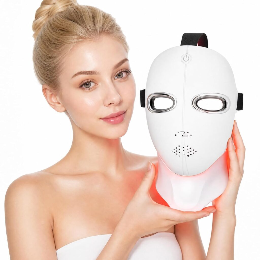A woman with blonde hair in a bun holds a white LED facial mask with silver eye areas and chin lights, smiling against a plain background.