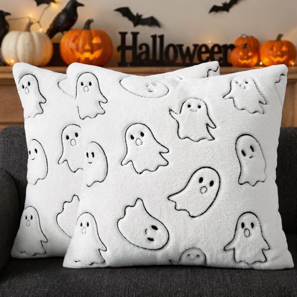 Two white Halloween Ghost Pillow Covers with cute, cartoon ghost designs sit on a dark couch. In the background are pumpkins, Halloween decorations, and black bat cutouts on the wall.