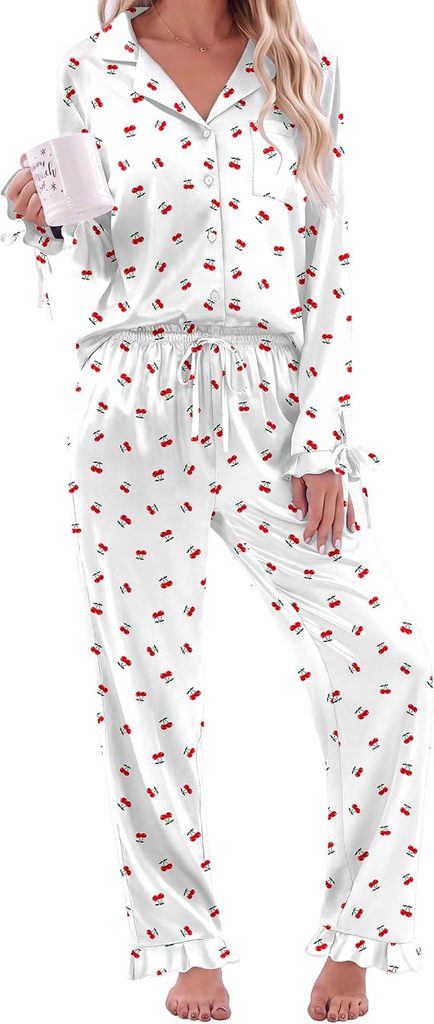 Woman wearing white pajama set with small red cherry prints, holding a mug in her right hand. The pajama top has buttons and the pants have a drawstring waist. She is barefoot and has long wavy hair.