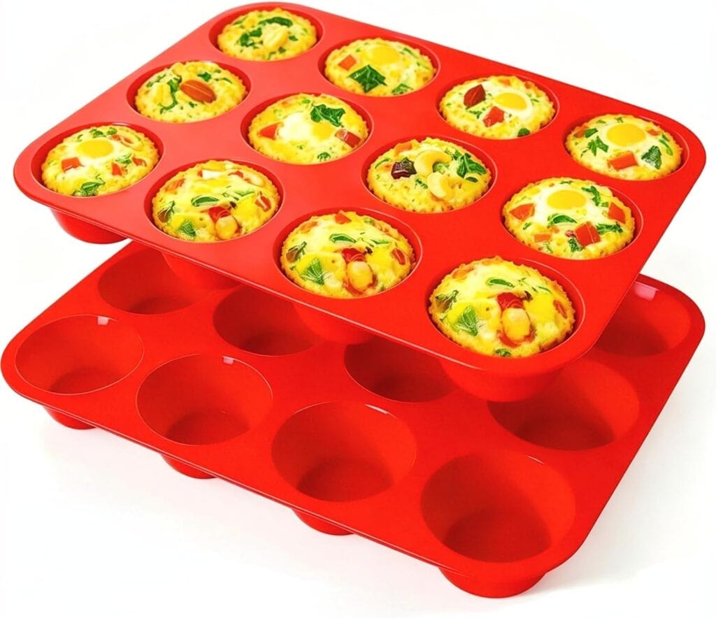 Two red silicone muffin trays, one filled with colorful baked egg muffins featuring veggies, and one empty, stacked at an angle on a white background.