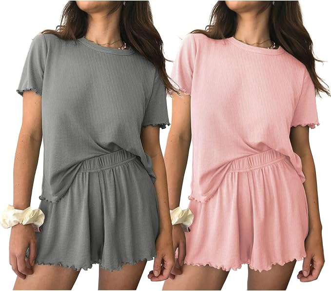 Two women wear matching ribbed short-sleeve lounge sets with elastic shorts. One set is gray, and the other is light pink. Both have a relaxed fit and wavy hems. Each woman wears a scrunchie on her wrist.