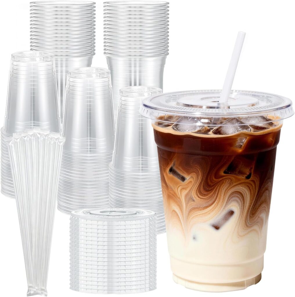 Clear plastic cups, lids, and straws are stacked in the background. In the foreground, an iced coffee with milk swirls sits in a clear cup with a lid and straw.