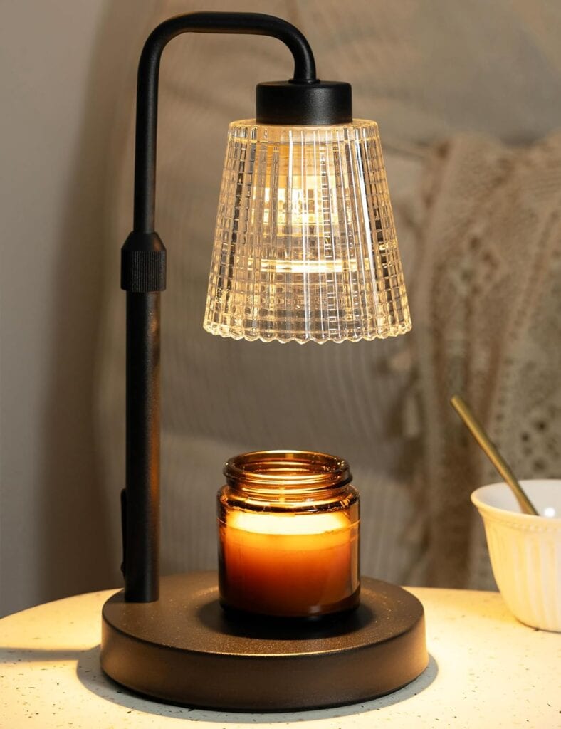 A black lamp with a glass shade hangs over a brown candle jar on a round table. In the background, there is a white knit blanket and part of a white bowl with a spoon.
