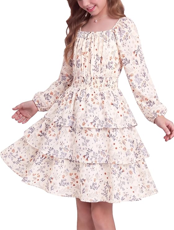 Young girl wearing a long-sleeved, ruffled, tiered dress with a floral pattern in soft colors. The dress has a gathered waist and a flowing skirt. She stands against a white background, smiling.