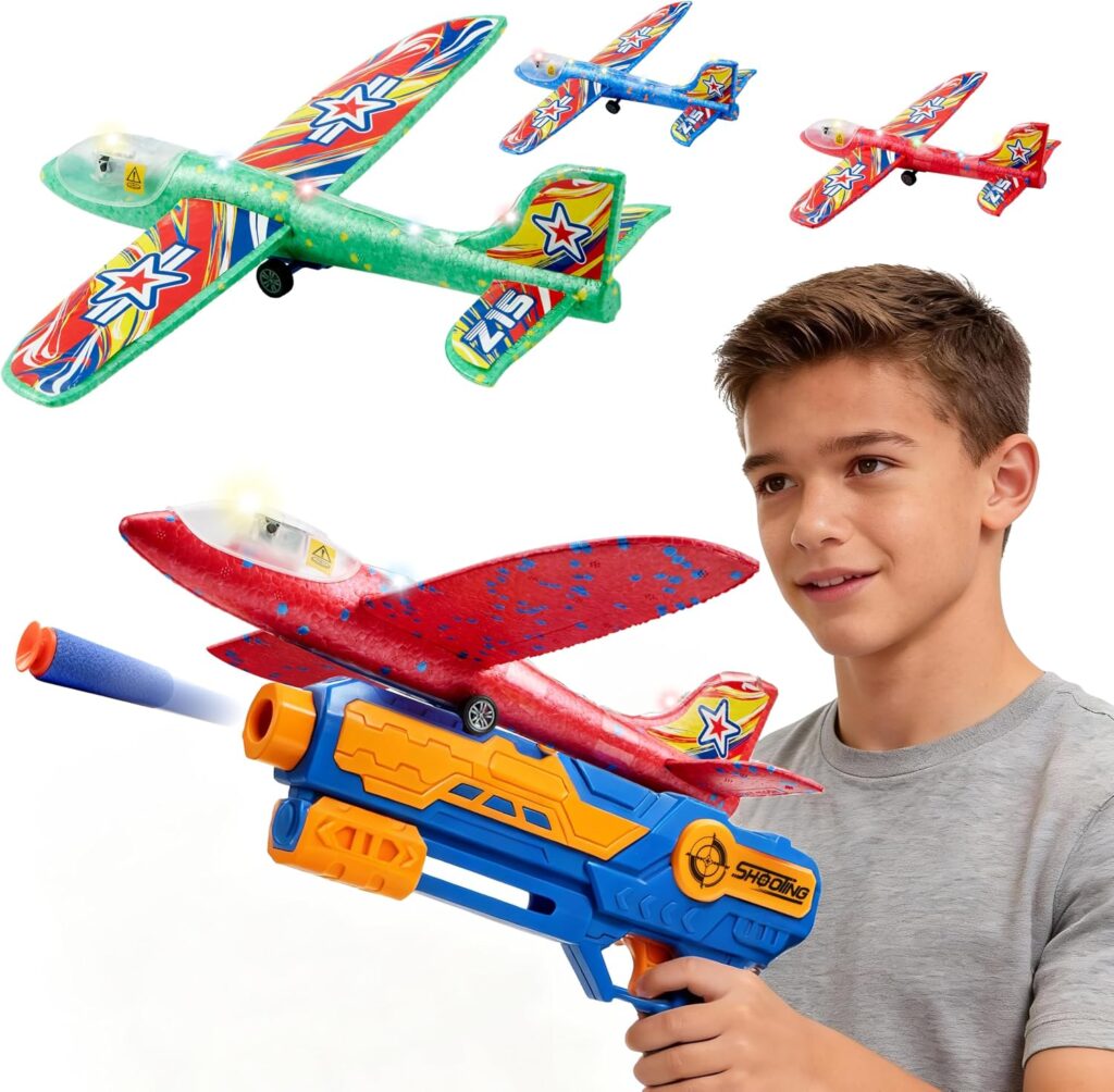 A boy holds a blue and orange toy launcher with a red foam glider airplane. Three colorful foam glider planes are flying in the background. The boy is smiling and looking at the glider.