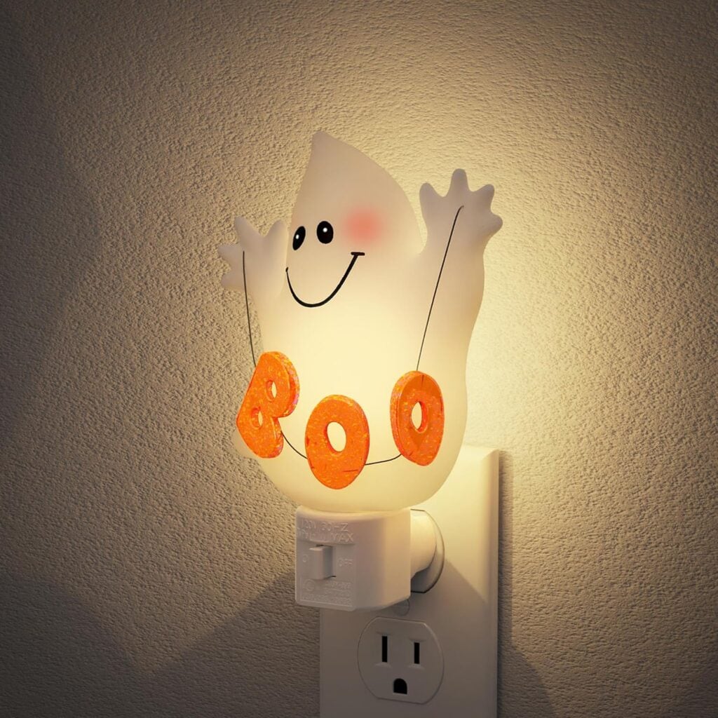A glowing nightlight shaped like a smiling ghost holding orange letters that spell “BOO,” plugged into a wall outlet against a beige textured wall.